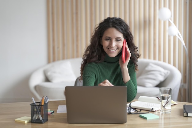 Happy Italian businesswoman having pleasant cellphone conversation while working on laptop at home