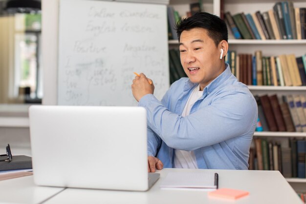 Счастливый японский учитель преподает онлайн, разговаривая с ноутбуком на рабочем месте