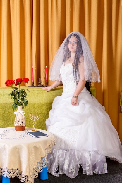 Una felice sposa ebrea in un abito bianco lussureggiante, il viso coperto da un velo, si trova nella stanza prima della cerimonia della chuppah. foto verticale