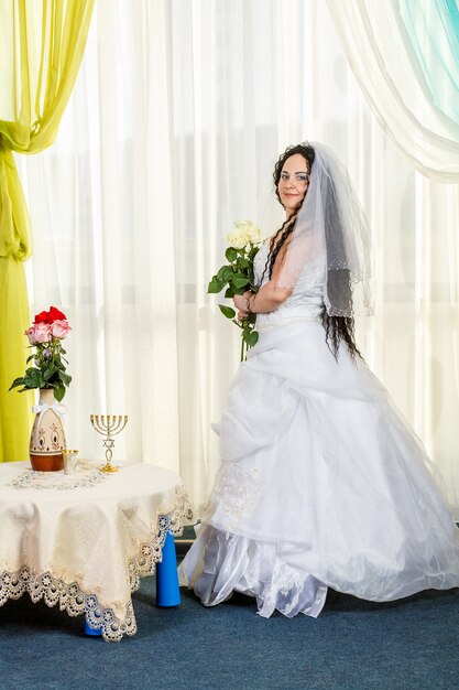 A happy Jewish bride stands in the hall before the chuppa ceremony at a table with flowers with a bouquet of white roses in her hands