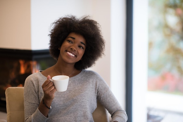 幸せな楽しいアフリカ系アメリカ人の女性は、暖炉でリラックスしながらコーヒーを飲みます。温かい飲み物を温めてウォーミングアップを持つ若い黒人の女の子。我が家の秋。