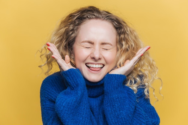Фото Счастливая радостная кудрявая женщина со здоровой кожей громко смеется держит глаза закрытыми в приподнятом настроении слышит смешные шутки от собеседника носит синий свитер позирует на фоне желтой студии