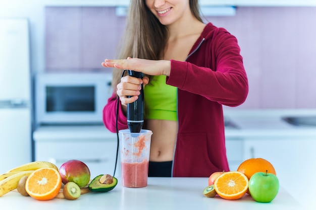 운동복에 행복 즐거운 맞는 여자는 부엌에서 집에서 핸드 블렌더를 사용하여 신선한 유기농 과일 스무디를 준비합니다. 건강한 식습관을위한 비타민 다이어트 음료