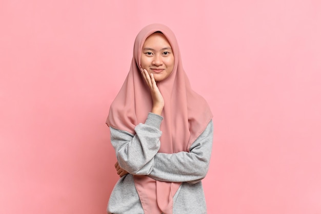 Happy and joyful look of  Young Asian Muslim woman with beautiful smile touch her cheek wears a pink veil and gray shirt on a pink background