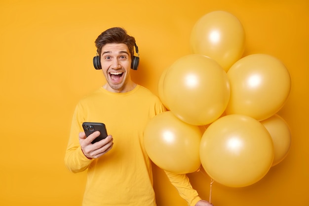 Happy joyful man listens music from playlist uses modern cellphone and wireless headphones wears casual jumper holds inflated balloons enjoys celebration isolated over vivid yellow background