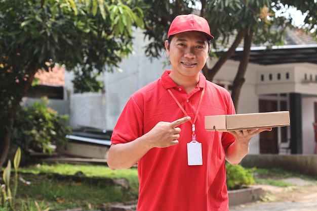 Foto uomo gioioso felice in uniforme rossa che sorride e che tiene la scatola di pizza all'aperto