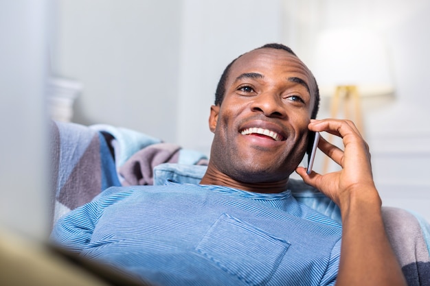 Felice gioioso uomo positivo sdraiato sul divano e sorridente mentre parla al telefono