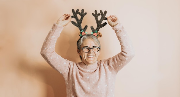 Счастливая радостная старшая старая женщина с белыми волосами в шляпе с оленьими рогами, улыбаясь во время игры и веселья. стоит на цветном фоне. В рождественской одежде. Скопируйте пространство. Место для рекламы