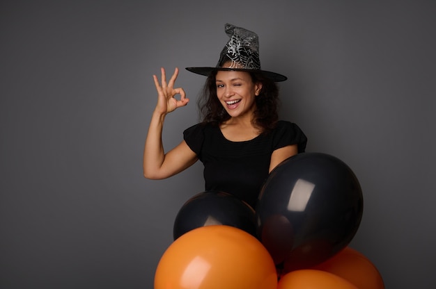 Happy joyful smiling toothy smile Hispanic woman wearing wizard hat, dressed in witch carnival costume for Halloween party, shows OK sign, poses against gray background with black balloons, copy space