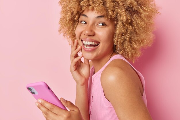 Happy joyful woman with curly hair laughs sincerely smiles
broadly uses mobile phone application dressed in t shirt isolated
over pink background cellular technology and online
communication