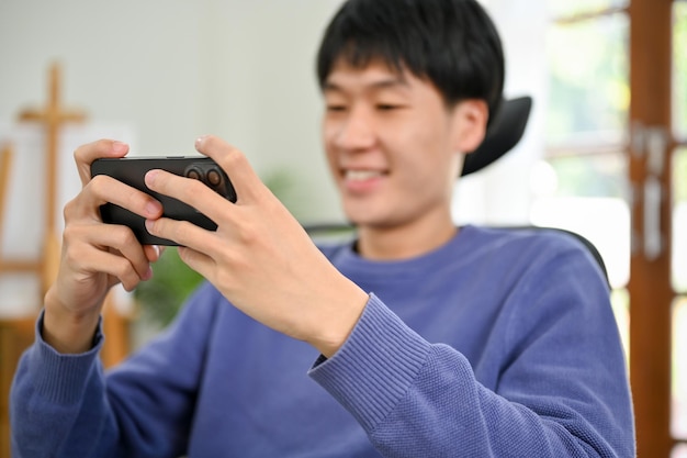 幸せでうれしそうな若いアジア人男性は、スマートフォンでモバイルゲームを楽しんでいます