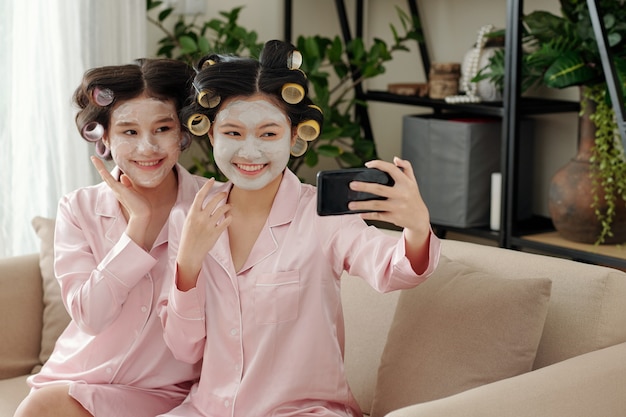 Happy joyful young vietnamese women taking selfie after applying clay mask on faces for moisturizing...