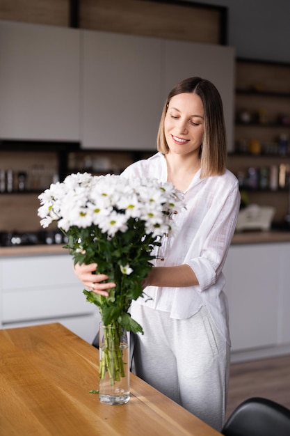 Felice e gioiosa giovane donna in bianco che dispone i fiori bianchi a casa in cucina