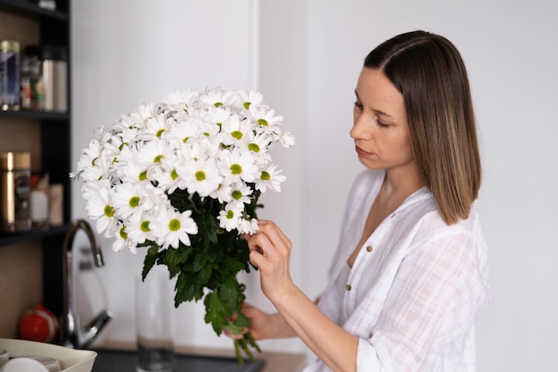 Foto felice e gioiosa giovane donna in bianco che dispone i fiori bianchi a casa in cucina