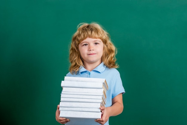 Bambino felice contro la lavagna verde concetto di istruzione e creatività bambino che tiene pila di libri con bordo di mortaio sulla lavagna ritorno a scuola scolaro in classe