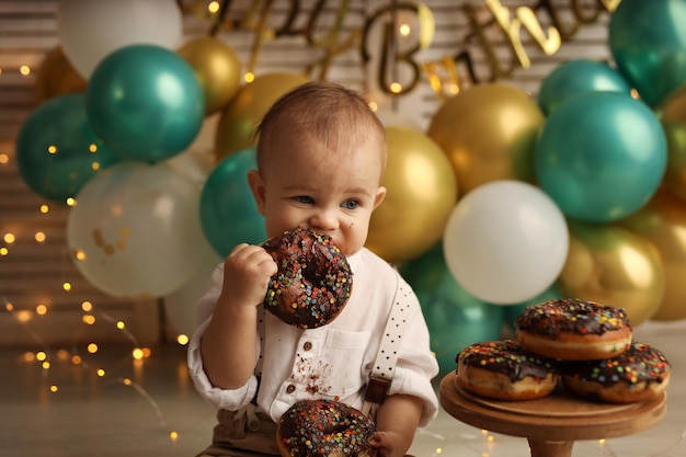 A happy kid on the background of balloons with garlands eats chocolate donuts