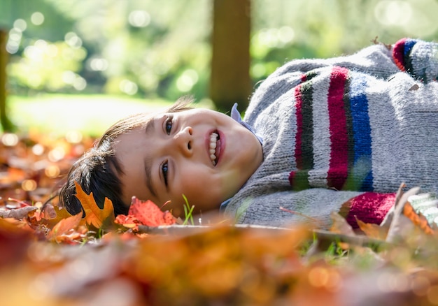 Happy kid boy laying down on leaves falling under the tree looking up at sky with smiling face