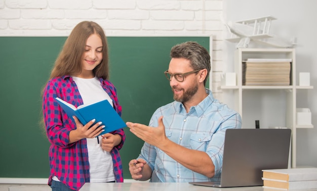 Happy kid and dad sitting in classroom with copybook and computer at blackboard education online