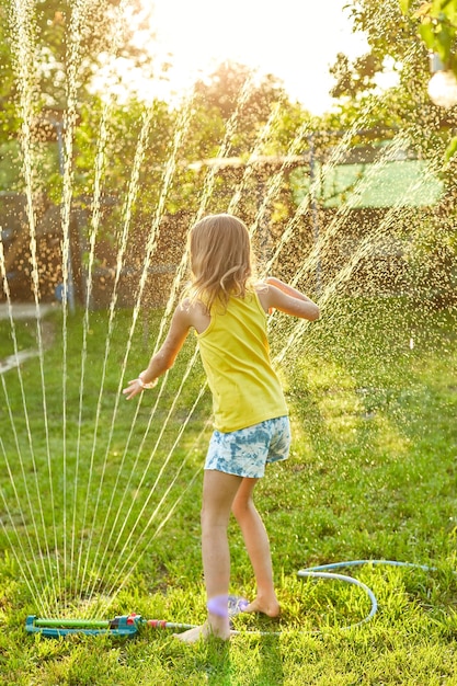 Счастливая девочка, играющая с садовым разбрызгивателем, бегает и прыгает летом