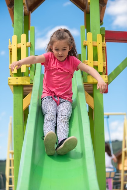 Happy kid girl riding on a slide at the playground while playing in the summer park