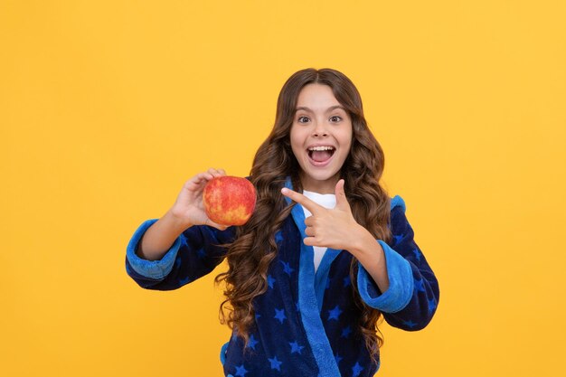 Happy kid girl in sleepwear pointing finger on fresh apple fruit, natural eating.