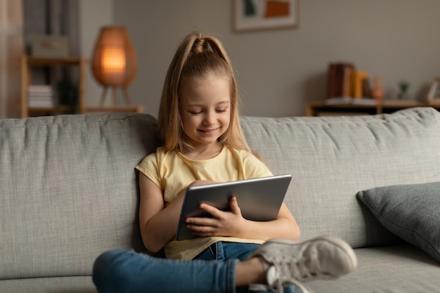 Happy Kid Girl Using Digital Tablet Sitting On Couch Indoor