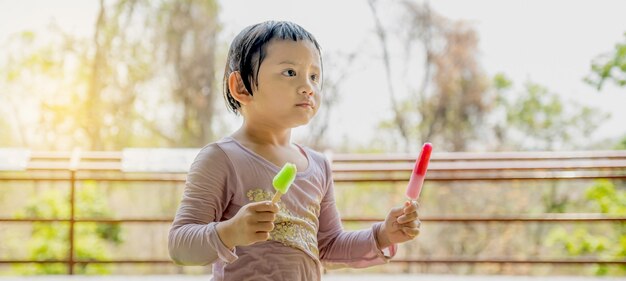 自然な屋外の背景でアイスクリームのアイスキャンディーを食べる濡れたシャツを着た幸せな子供の女の子。