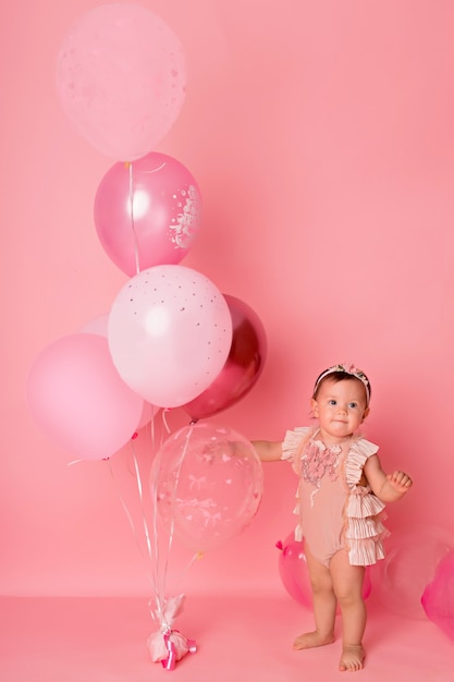 Happy kid girl with balloons on a pink background celebrates her first birthday