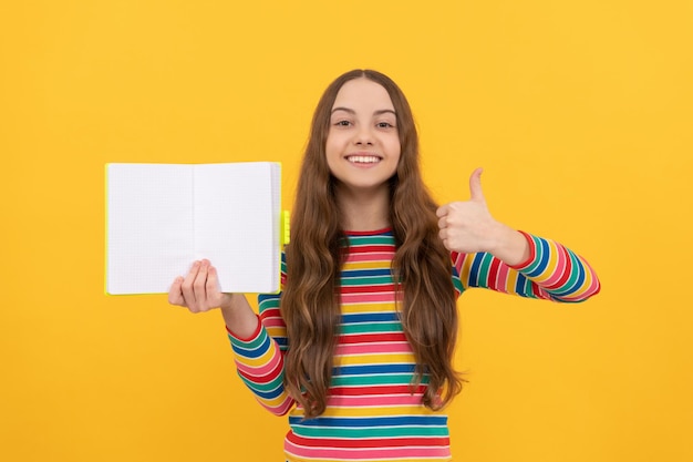 Happy kid give thumbs up hand gesture holding open book for copy space yellow background, satisfied.