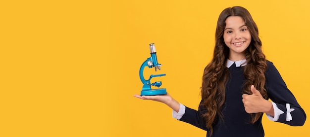 Happy kid hold microscope for school education on yellow background showing thumb up biology horizontal isolated poster of school girl student banner header portrait of schoolgirl copy space