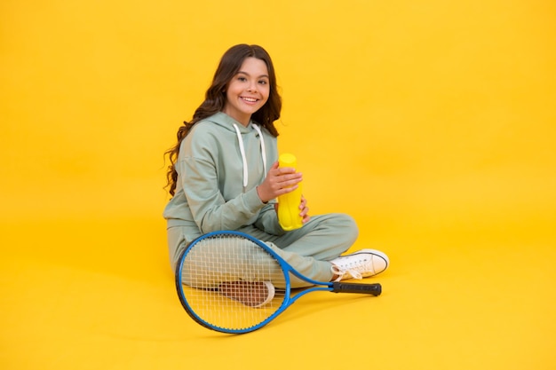 Happy kid hold tennis racket and drink water from bottle on\
yellow background, hydration.