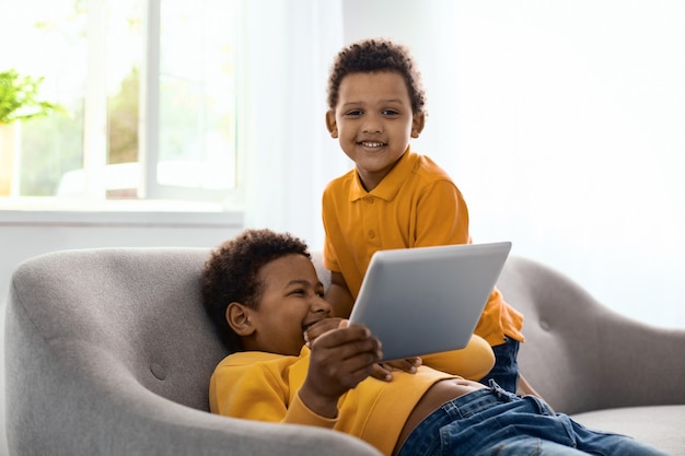 Счастливый ребенок. Радостный маленький мальчик позирует перед камерой и улыбается, глядя на мультфильм со своим братом на планшете и щекоча его