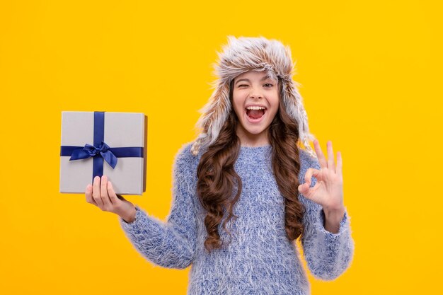 Happy kid long hair in hat with gift box show ok on yellow background holiday