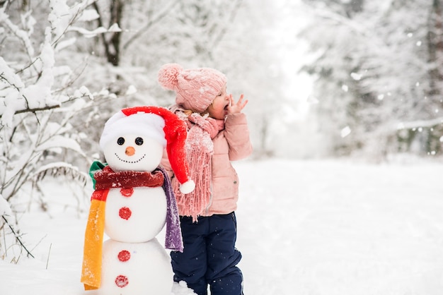 雪だるまと遊ぶ幸せな子供。屋外の冬の散歩に面白い小さな女の子