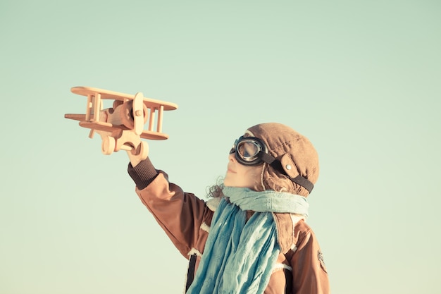 Happy kid playing with toy wooden airplane against autumn sky background. Retro toned