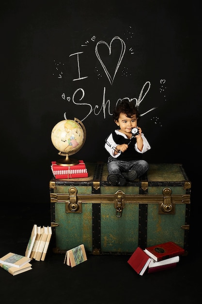 Happy kid preschool boy with books, globe and clock near school board blackboard sitting on a chest