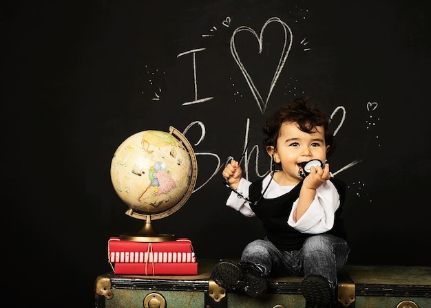 Happy kid preschool boy with books, globe and clock near school board blackboard sitting on a chest