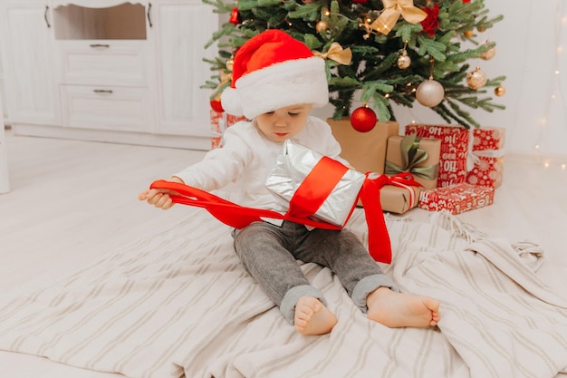 A happy kid in a Santa hat sits on the floor near the Christmas tree and holds a box with a gift in his hands.