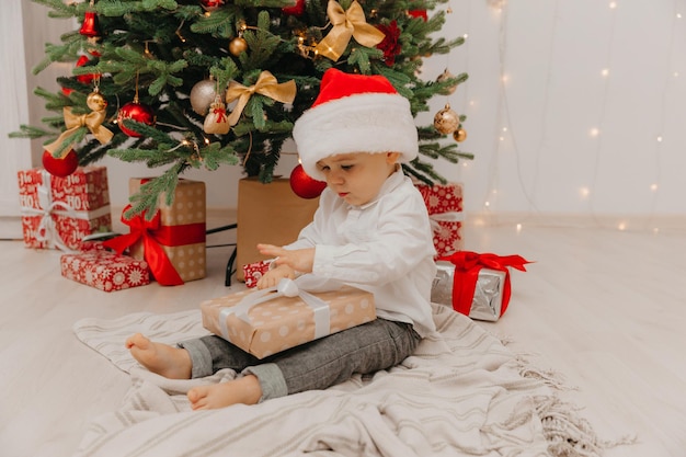 A happy kid in a Santa hat sits on the floor near the Christmas tree and holds a box with a gift in his hands.