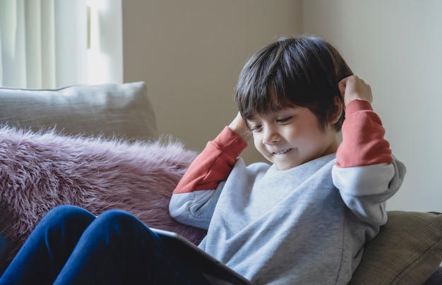 ソファの上に座って幸せな子供はタブレットで彼のクラスの友達と一緒に取って楽しいデジタルタブレットでオンラインで勉強している子供の少年