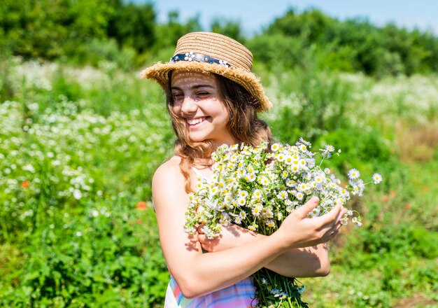 Bambino felice sorriso in cappello da sole con mazzo di fiori di camomilla in vacanza paesaggio soleggiato in campagna estate estate