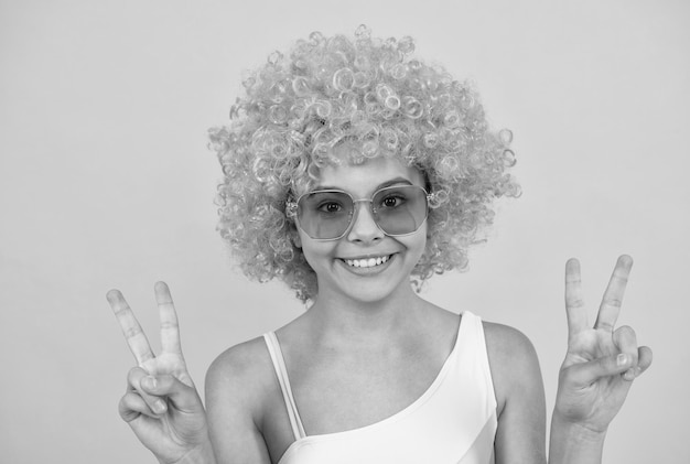 Happy kid in sunglasses and swimsuit wearing orange wig with peace gesture on yellow background fun