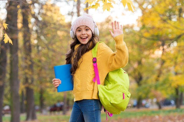 happy kid wear headphones in autumn park with book and backpack school education