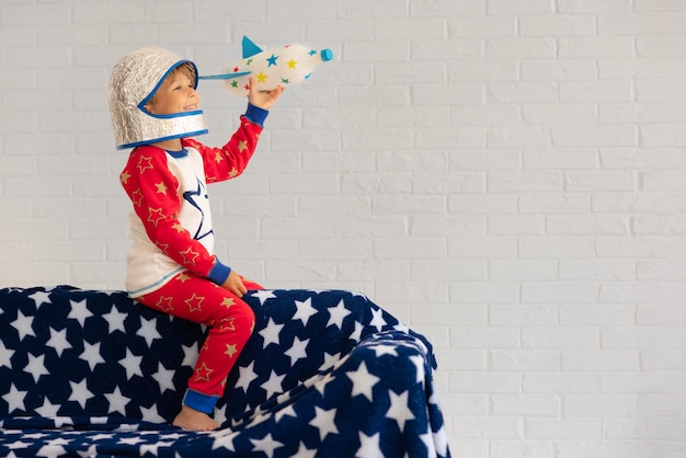 Happy kid with craft toy rocket. Child playing at home against white wall background. Kid pretend to be astronaut. Imagination and children dream concept