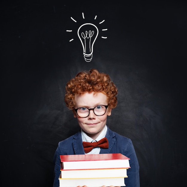 Happy kid with ginger hair holding stacks of book on school blackboard background with idea lightbulb