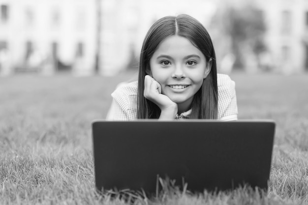 Счастливый ребенок, работающий на ноутбуке, онлайн-обучение обратно в школу. Девушка-подросток использует компьютер на зеленой траве в парке. Ребенок с ноутбуком. Новые технологии в современной жизни.