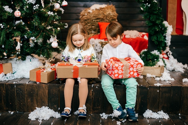 Happy kids opening gift boxes in studio with cristmas tree and new year decorations.