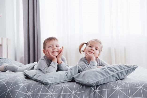 Happy kids playing in white bedroom. Little boy and girl, brother and sister play on the bed wearing pajamas. Nursery interior for children. Nightwear and bedding for baby and toddler. Family at home