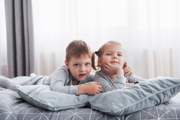 白い寝室で遊んでいる幸せな子供たち。男の子と女の子、弟と妹はパジャマを着てベッドで遊ぶ。子供のための保育園のインテリア。ベビーと幼児用のナイトウェアと寝具。自宅で家族