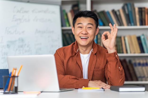 Happy Korean Teacher At Laptop Gesturing Okay Sitting At Workplace